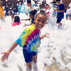 A smiling girl in a tie-dye t-shirt plays in a mountain of bubble with other boys and girls play together in the background