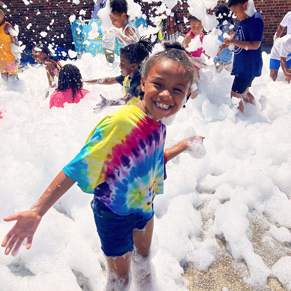A smiling girl in a tie-dye t-shirt plays in a mountain of bubble with other boys and girls play together in the background